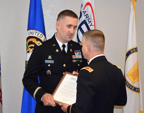 Two soldiers in dress uniforms exchange charter in front of flags.