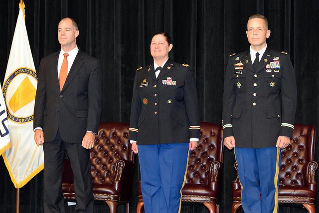 Three people stand on stage in front of chairs.